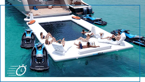 Aerial view of a large FunAir Beach Club Sea Pool with guests lounging on inflatable deck areas, jet skis surrounding it in turquoise tropical water