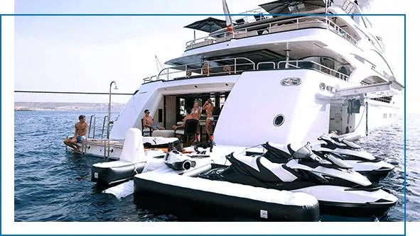 Stern view of a large white superyacht with multiple jet skis docked on a custom inflatable platform, guests visible on aft deck, Mediterranean coastline in background