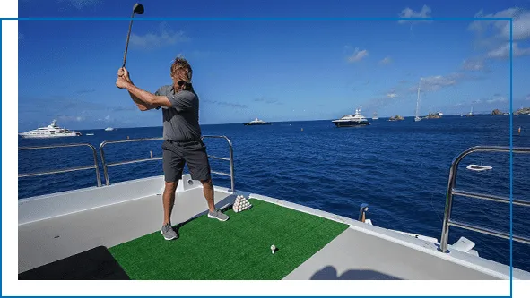 Man playing golf from the deck of a superyacht towards floating inflatable golf greens, other yachts anchored in the background