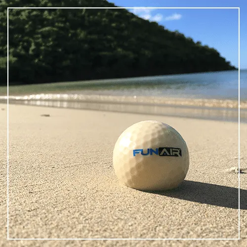 FunAir branded biodegradable golf ball resting on a sandy beach with tropical hillside in background